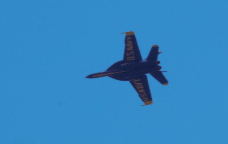 An F/A-18 Super Hornet E/F painted in Blue Angels colors tears through the sky above my neighborhood. On the underside, “US Navy” is emblazoned in yellow.